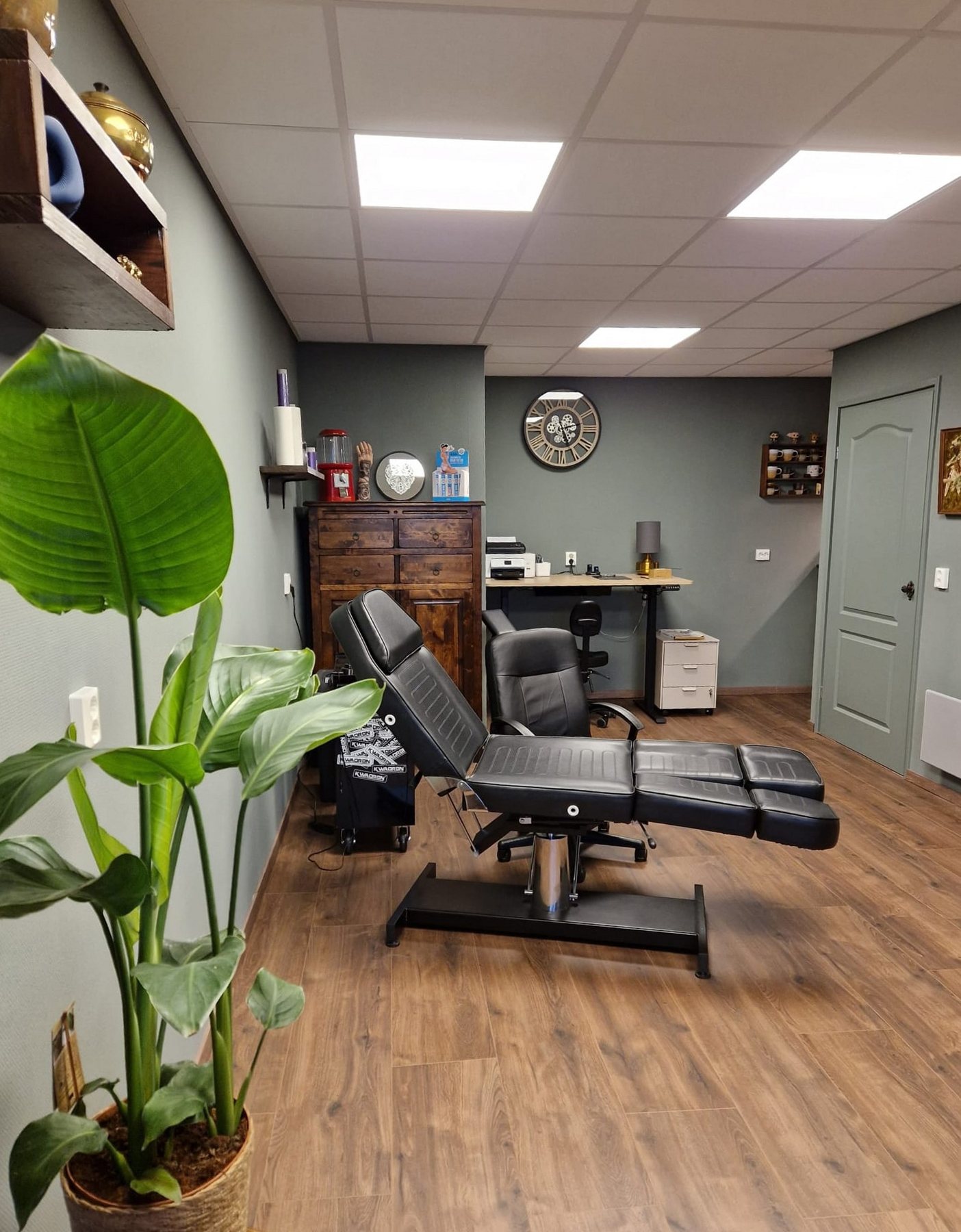 De nieuwe studio: zwarte tattoostoel, bureau, klok aan de grijze muur en een grote plant